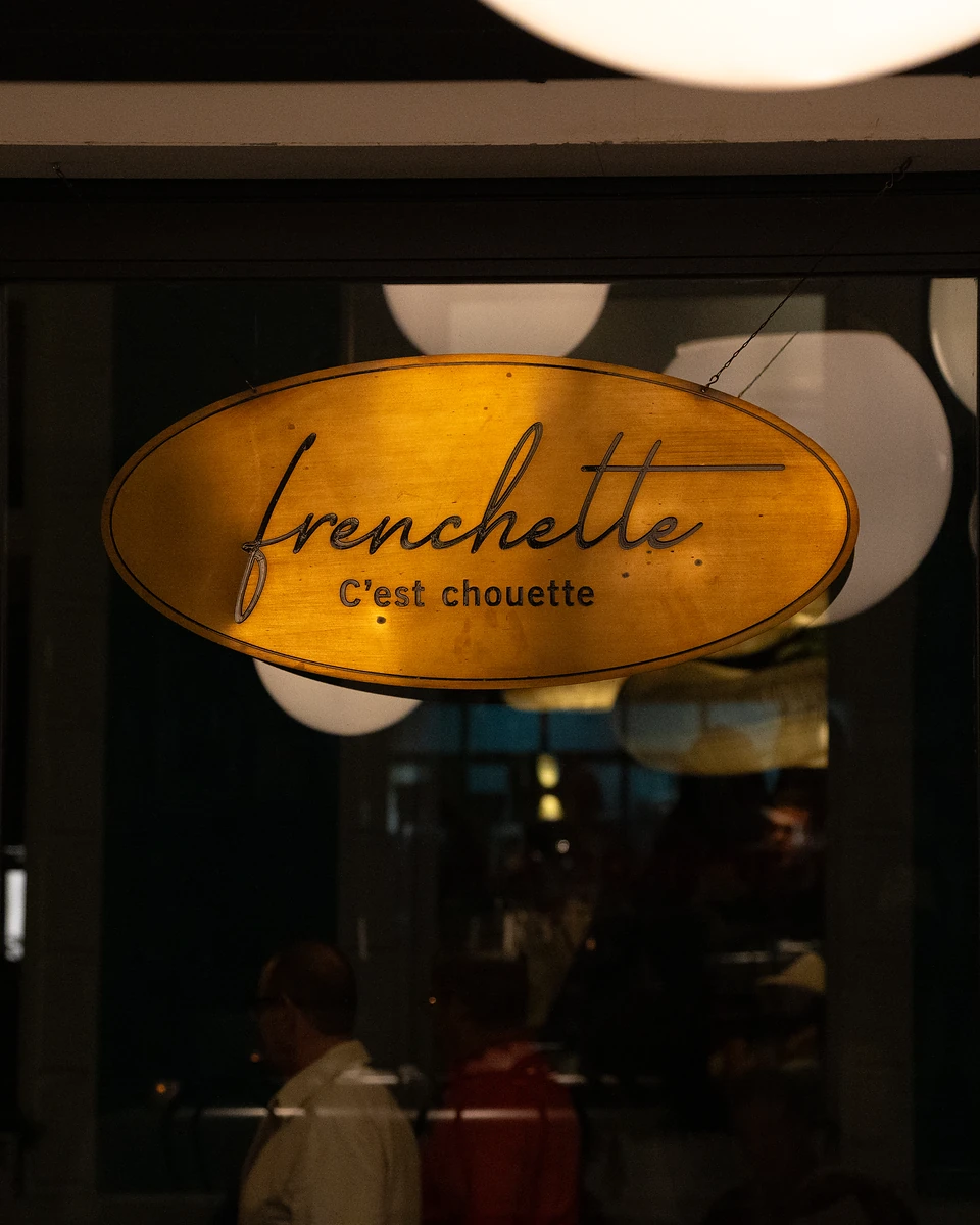 The golden oval sign reading 'frenchette, C'est chouette' in the window, globe lamps glowing behind