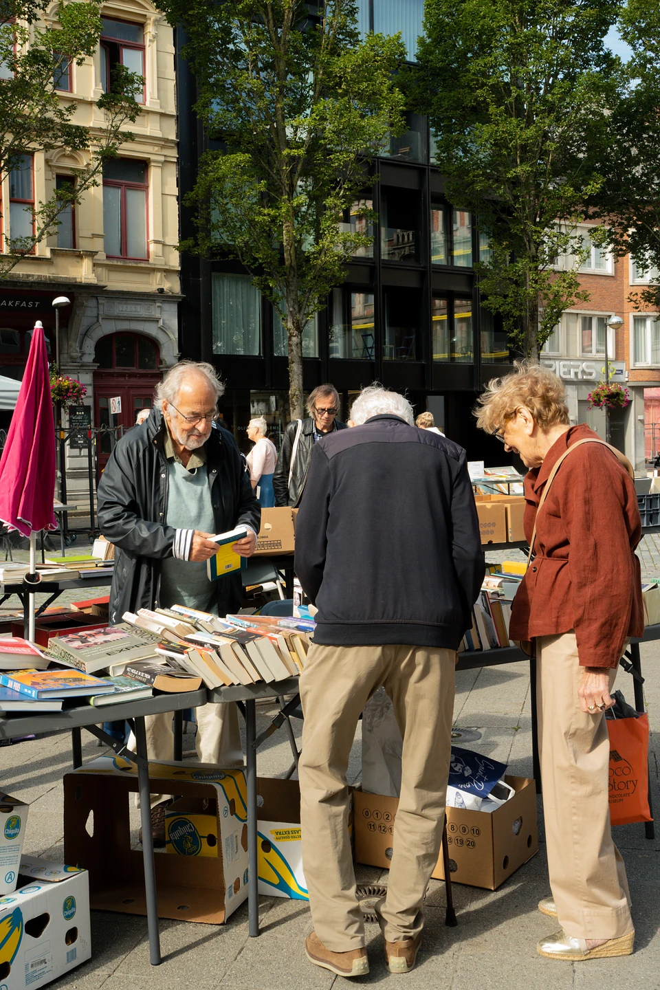 People browsing tables and boxes of books at an open-air book stall under trees