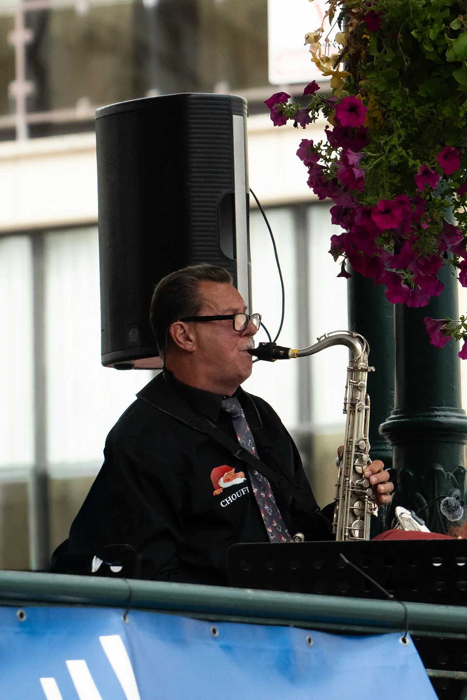 A saxophonist in a black shirt and tie playing in the kiosk beside hanging pink flowers