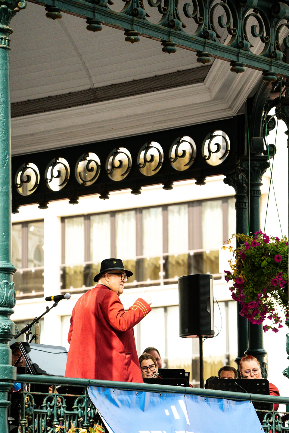 A conductor in a red coat and black hat leading the band in the Wapenplein kiosk