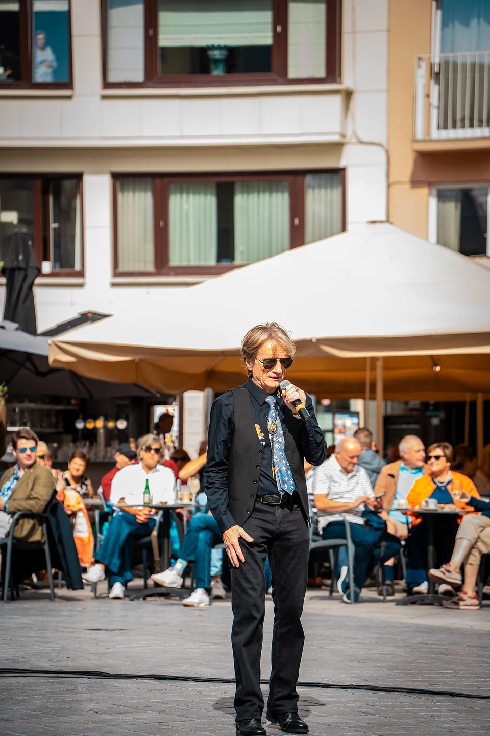 A singer in sunglasses and a black waistcoat performing on the square, café terraces behind